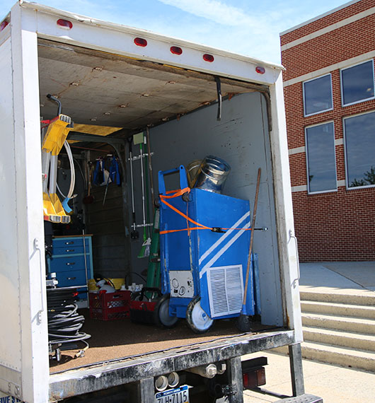 A moving truck parked near a building, with its rear door open revealing an assortment of equipment and bins for mold removal, secured with orange straps.